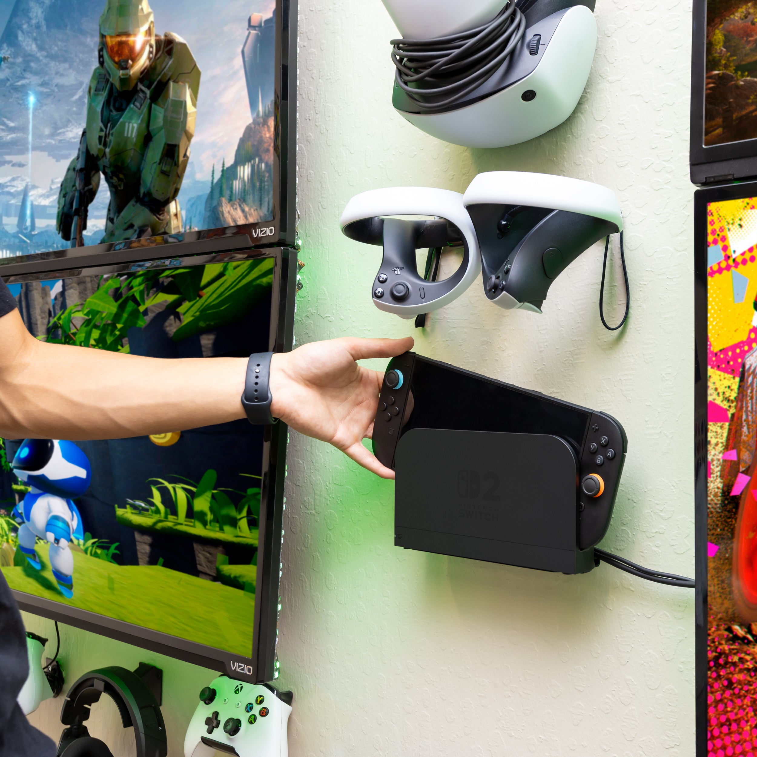 Gamer placing Nintendo Switch 2 console into a black HIDEit Switch 2 wall mount, surrounded by dual monitors, VR headset, and gaming accessories in a modern gaming setup.